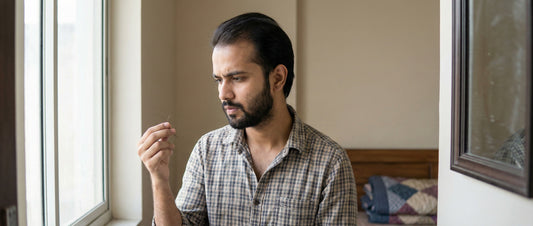 Young Indian man in his 20s examining mild hair thinning near a window, reflecting uncertainty about whether biotin can realistically help early hair fall.